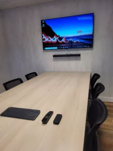 Salle de réunion moderne équipée d'un écran de présentation mural, table de conférence en bois clair, système audiovisuel intégré et télécommandes pour visioconférence